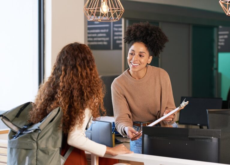 Young,Woman,Checks,In,At,Front,Desk,,Reception,,Hotel,,College